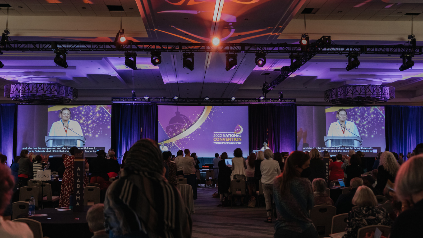 A photo of attendees in the ballroom at Convention 2022 in Denver, CO. The photographer is taking the picture from the back of the room, facing the stage. The crowd is featured in the image. The stage has three screens - the one in the middle has the Convention 2022 logo ("2022 National Convention: Women Power Democracy"). The two other screens on the right and left feature a LWVUS board member speaking on stage.